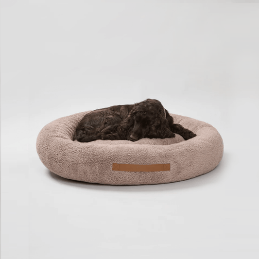 A brown round dog bed with a plush texture, featuring a lying dog resting on it.